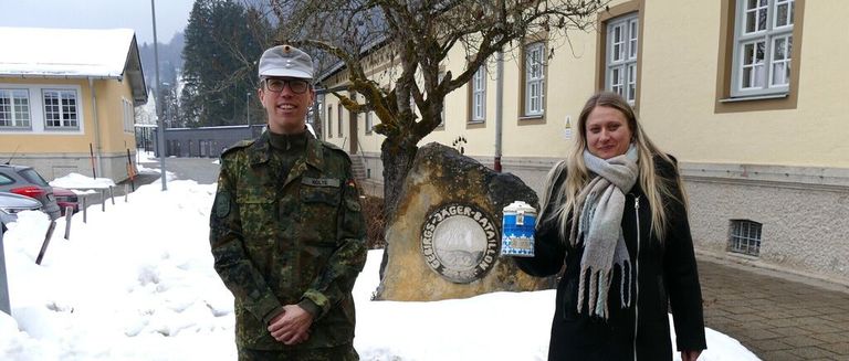 Ein Mann in Uniform und eine Frau in einem Mantel posieren vor einem Stein mit einem Emblem. Sie stehen vor einem Gebäude.