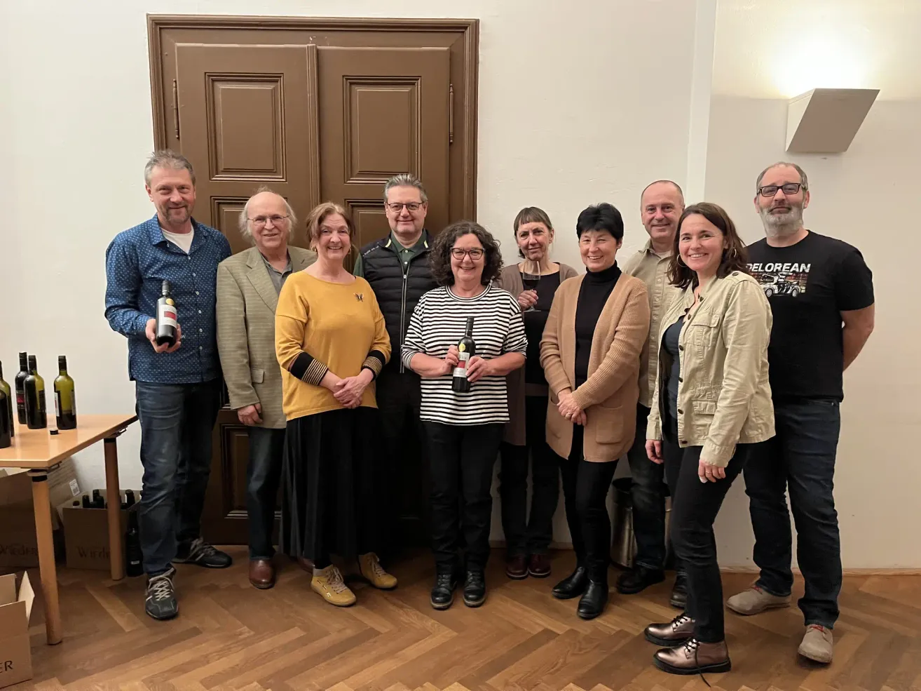 Eine Gruppe von Menschen, sowohl Männer als auch Frauen, stehen in einem Raum und halten Weinflaschen in der Hand, lächeln für ein Foto.