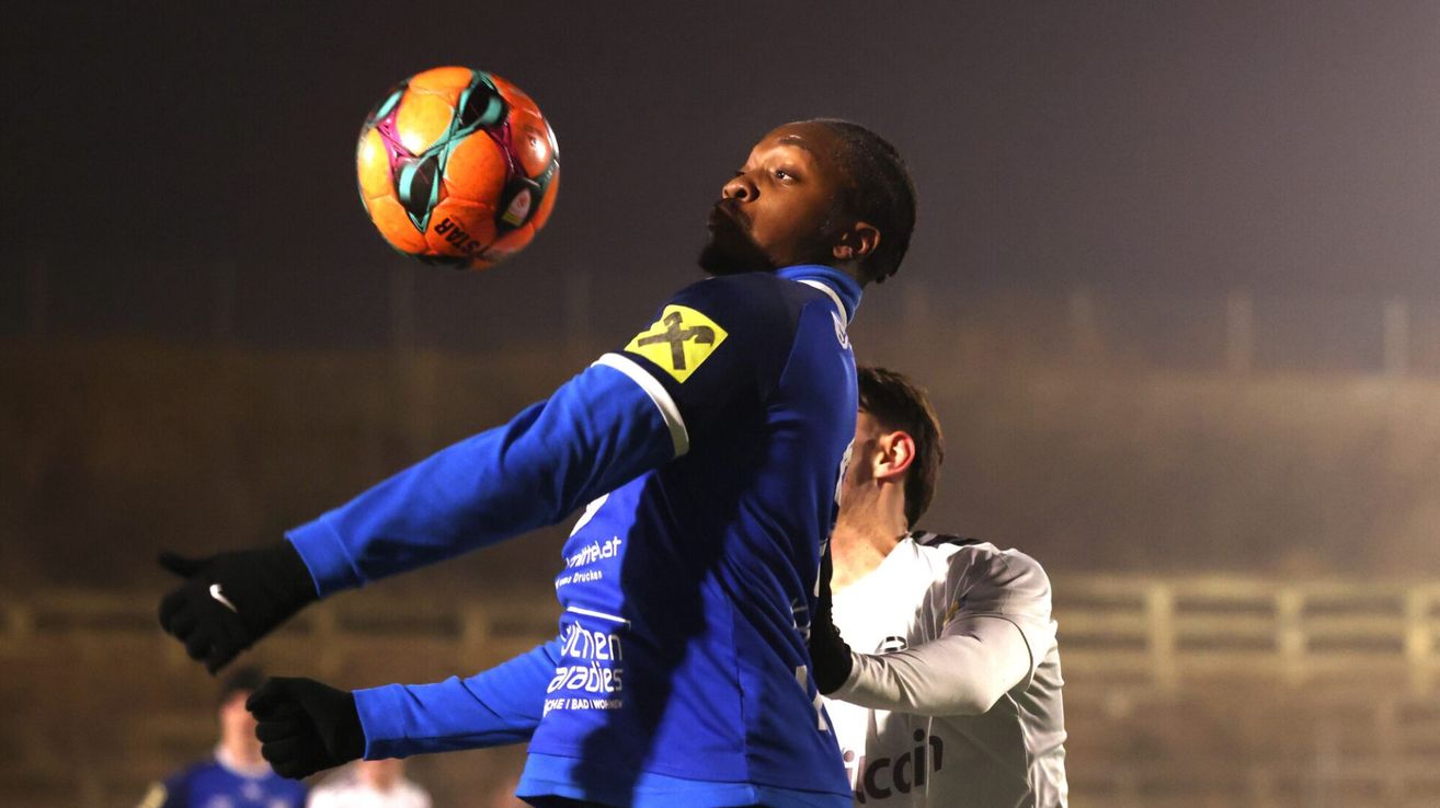 Ein Fußballspieler in einem blauen Trikot springt nach dem Ball in einem Nachtspiel. Hinter ihm steht ein weiterer Spieler in einem weißen Trikot.