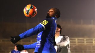 Ein Fußballspieler in einem blauen Trikot springt nach dem Ball in einem Nachtspiel. Hinter ihm steht ein weiterer Spieler in einem weißen Trikot.