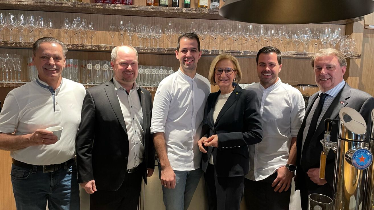 Vier Menschen stehen lächelnd vor einer Bar. Drei Männer tragen weiße Kochjacken, und eine Frau trägt einen schwarzen Anzug. Dahinter befinden sich Regale mit Weingläsern und Spirituosenflaschen.