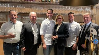 Vier Menschen stehen lächelnd vor einer Bar. Drei Männer tragen weiße Kochjacken, und eine Frau trägt einen schwarzen Anzug. Dahinter befinden sich Regale mit Weingläsern und Spirituosenflaschen.