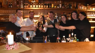 Eine Gruppe von Menschen feiert mit Weinglasern in einer Bar. Regale mit Flaschen und Gläsern stehen hinter ihnen.