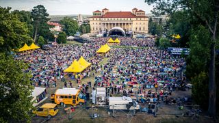 Eine große Menschenmenge ist im Freien versammelt, mit vielen aufgestellten Zelten. Eine Bühne ist im Hintergrund zu sehen. Fahrzeuge und temporäre Strukturen sind verstreut.