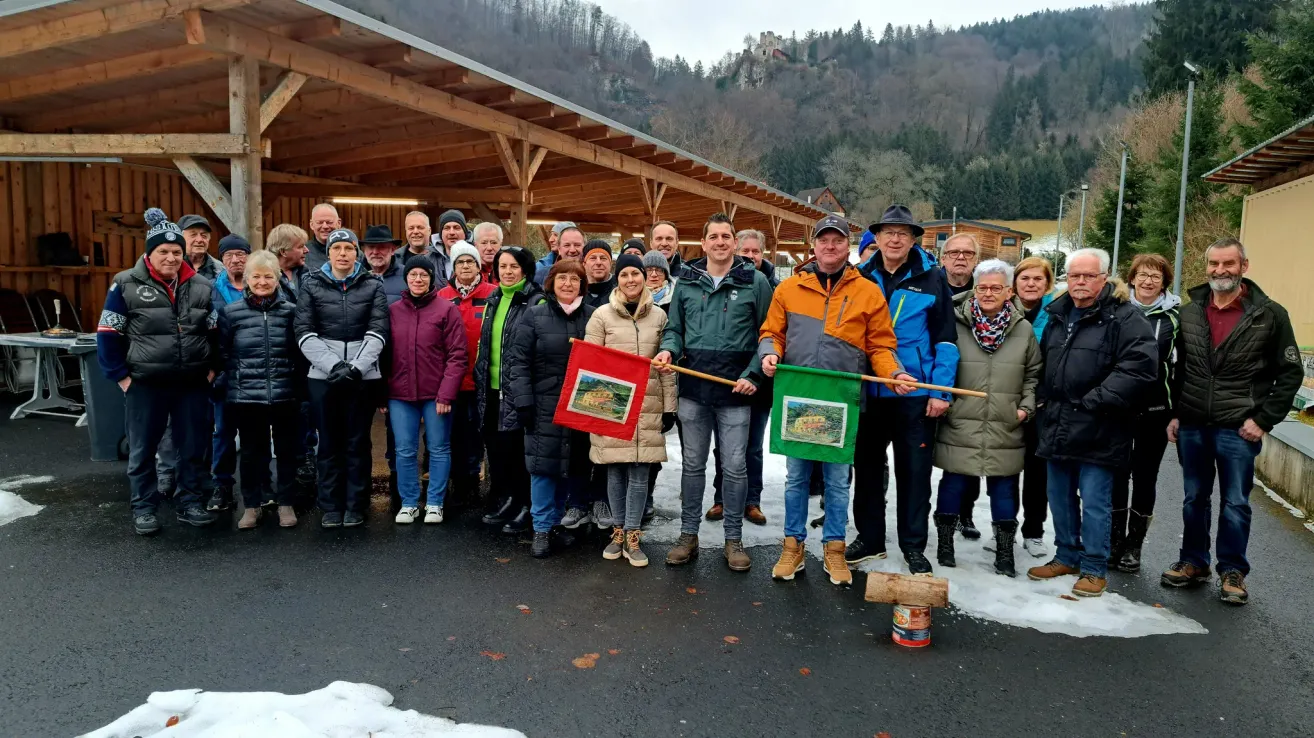 Eine Gruppe von Menschen steht draußen in einem verschneiten Bereich und trägt Winterkleidung. Sie stehen vor einer Holzkonstruktion. Zwei Personen halten Fahnen. Ein kleiner Kanister liegt auf dem Boden.