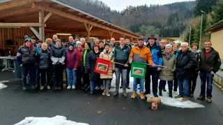 Eine Gruppe von Menschen steht draußen in einem verschneiten Bereich und trägt Winterkleidung. Sie stehen vor einer Holzkonstruktion. Zwei Personen halten Fahnen. Ein kleiner Kanister liegt auf dem Boden.
