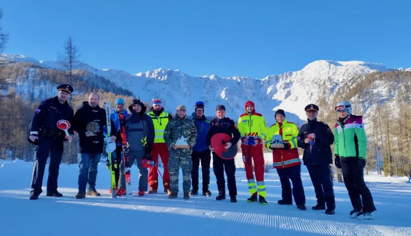 Eine Gruppe von Menschen steht im Schnee mit Bergen im Hintergrund, trägt Winterkleidung und hält Trophäen und Skier. Einige tragen Militäruniformen.