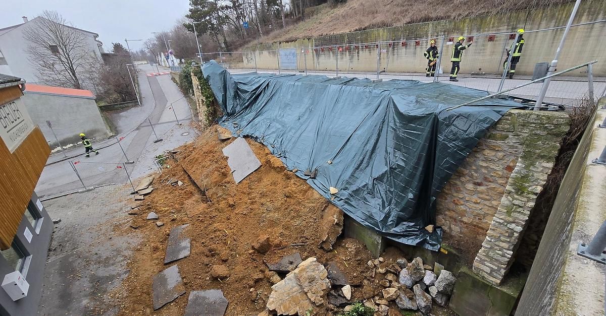 Eine Straße neben einer Brücke, die mit einer Plane abgedeckt ist. Zwei Feuerwehrleute stehen daneben. Schutt liegt auf dem Boden und eine Steinmauer ist in der Nähe.