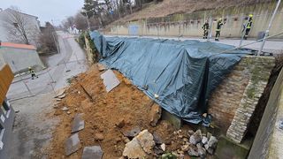 Eine Straße neben einer Brücke, die mit einer Plane abgedeckt ist. Zwei Feuerwehrleute stehen daneben. Schutt liegt auf dem Boden und eine Steinmauer ist in der Nähe.