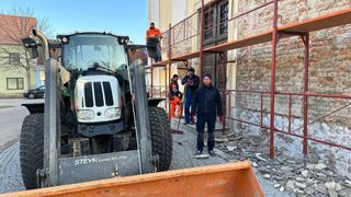Mehrere Arbeiter sind mit Renovierungsarbeiten außerhalb eines Gebäudes beschäftigt. Ein Traktor ist in der Nähe geparkt, und ein Mann geht auf das Gebäude zu.
