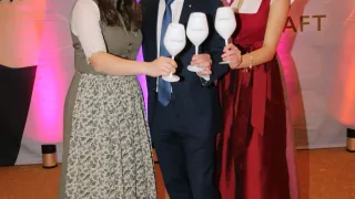 Drei Personen, zwei Frauen und ein Mann, gekleidet in formeller Kleidung und mit Weingläsern in den Händen, lächeln und posieren für ein Foto.