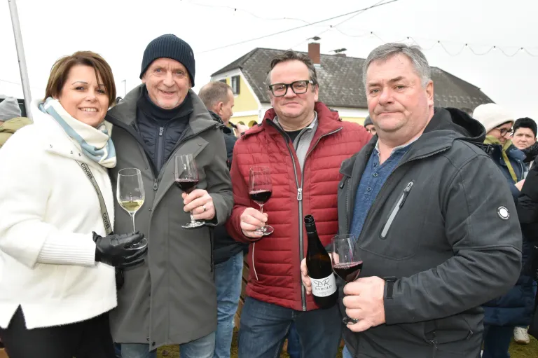 Vier Erwachsene lächeln, halten Weingläser in der Hand und posieren für ein Foto im Freien. Dahinter steht ein Haus und eine Straßenlaterne.