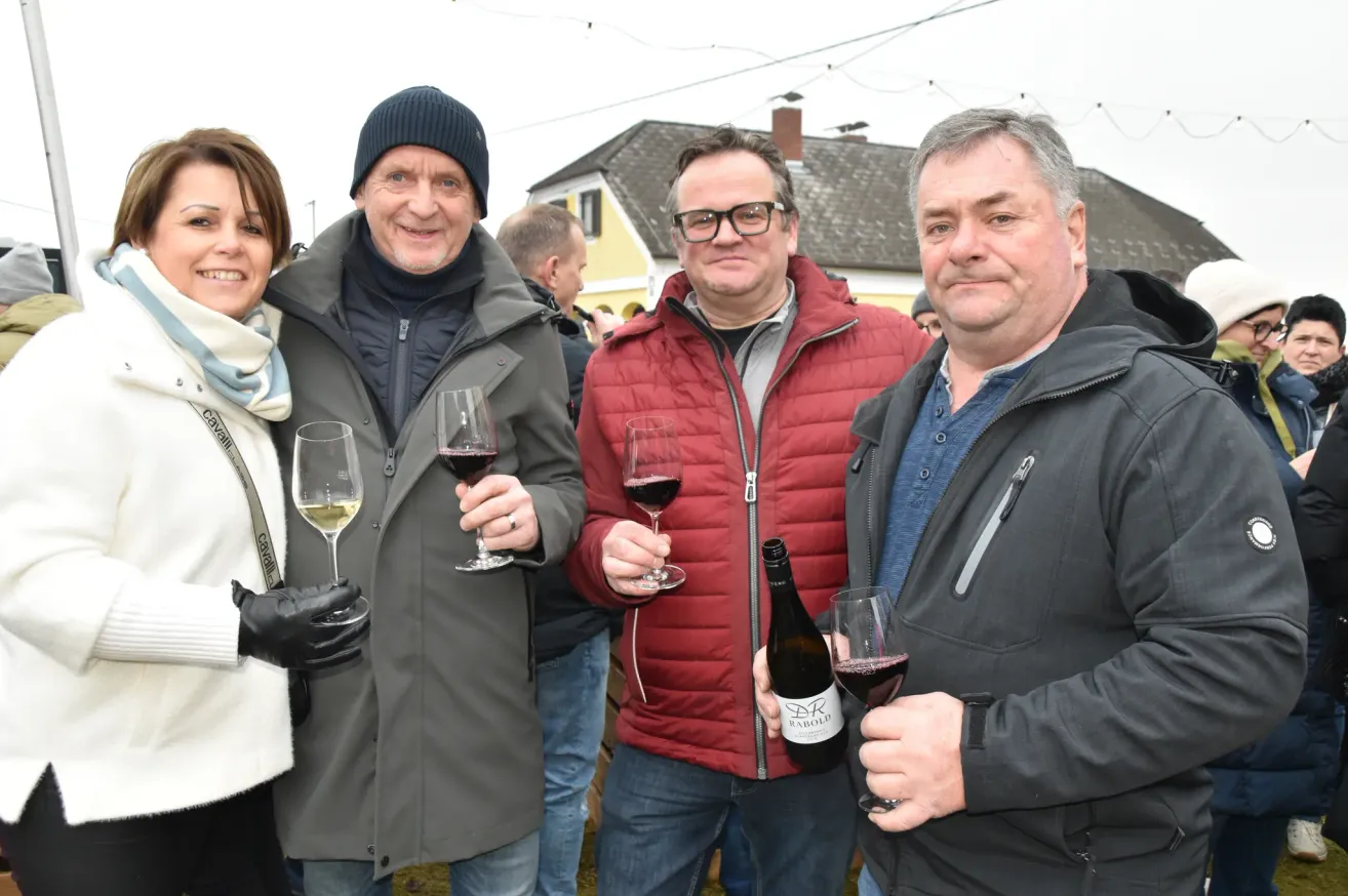 Vier Erwachsene lächeln, halten Weingläser in der Hand und posieren für ein Foto im Freien. Dahinter steht ein Haus und eine Straßenlaterne.