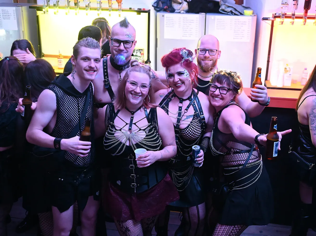 Eine Gruppe von Menschen in Netz- und Lederoutfits posiert mit Getränken für ein Foto in einer Bar.