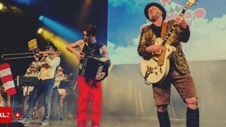 Eine Band tritt auf der Bühne auf mit einem Mann, der Gitarre spielt, einem anderen, der Akkordeon spielt, einem Trompeter und einem Schlagzeuger. Scheinwerfer und eine Leinwand im Hintergrund.