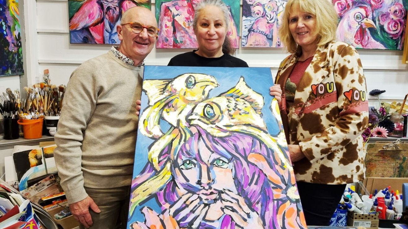 Drei Personen stehen neben einem großen Gemälde einer Frau mit lila Haaren und grünen Augen und halten es. Hinter ihnen hängen weitere Gemälde an der Wand.