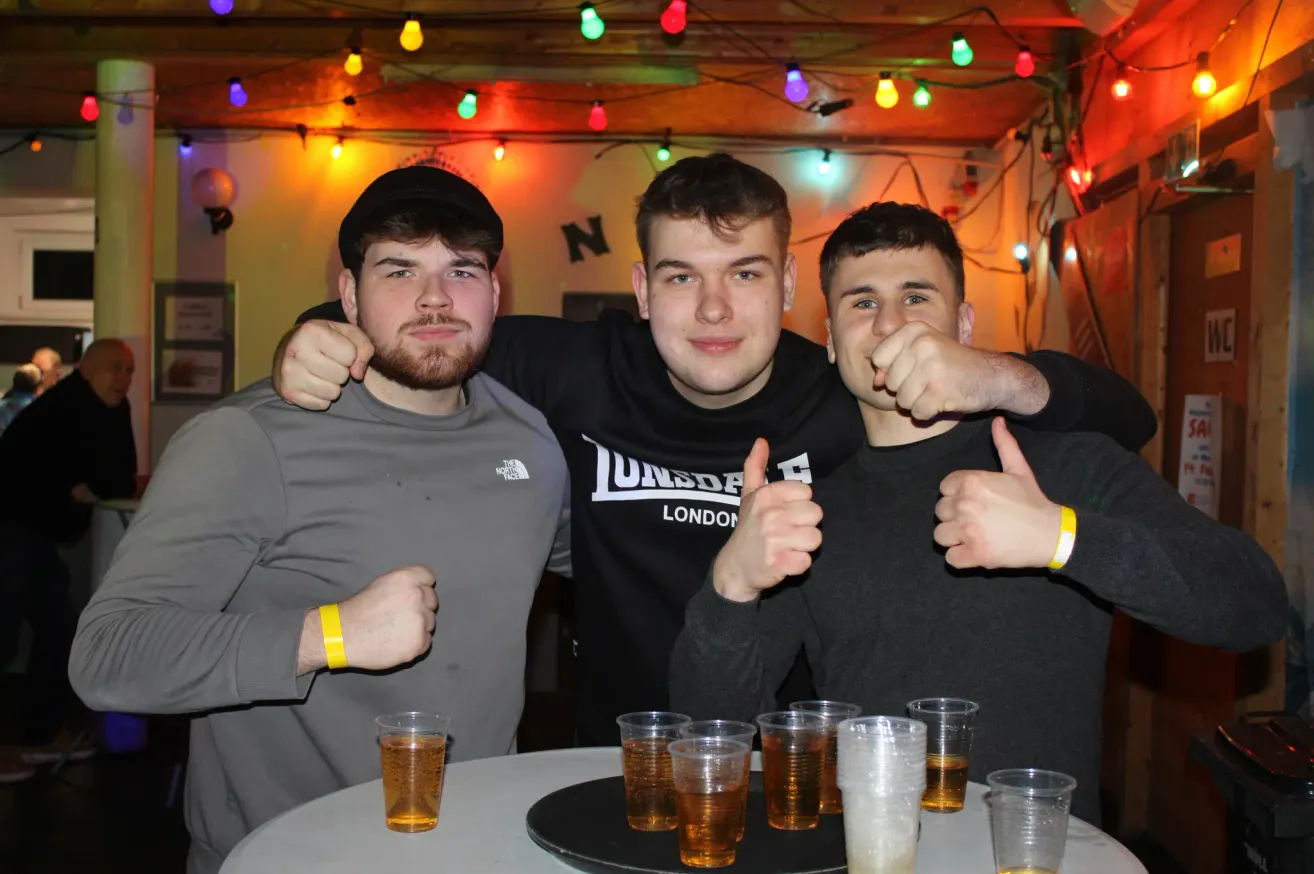Drei junge Männer mit Armbändern posieren für ein Foto in einer Bar mit Biergläsern auf einem Tisch. Sie sind von farbigen Weihnachtslichtern umgeben.