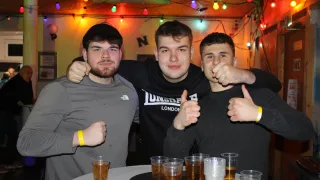 Drei junge Männer mit Armbändern posieren für ein Foto in einer Bar mit Biergläsern auf einem Tisch. Sie sind von farbigen Weihnachtslichtern umgeben.