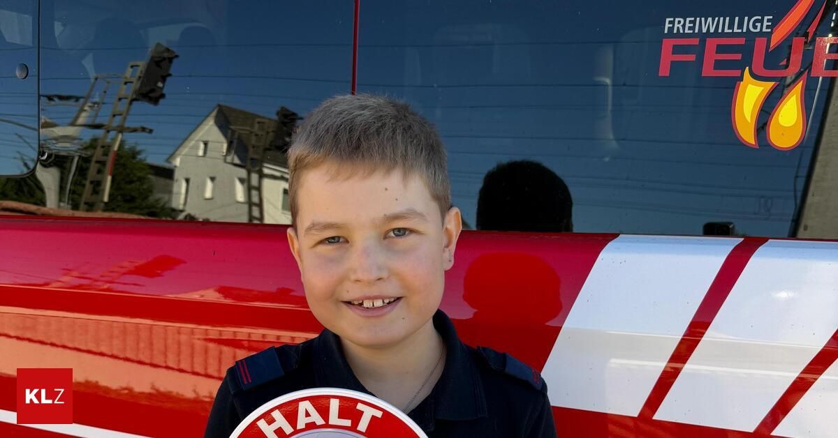Ein junger Junge trägt eine marineblaue Uniform mit dem Wort Halt auf einem kreisförmigen Schild, das er in der Hand hält. Er steht vor einem roten Fahrzeug.