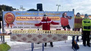 Ein Mann steht vor einem Tisch voller Trophäen, mit einem großen Banner hinter ihm, auf dem ein Skifahrer und die Website www.schisegger.at abgebildet sind.
