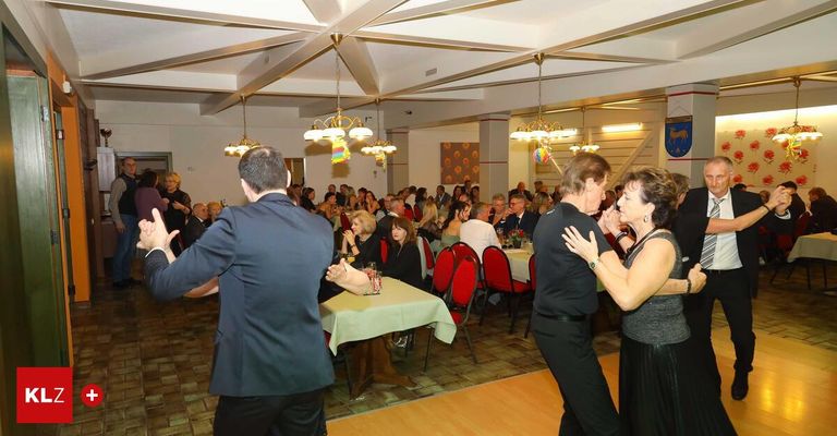 Eine Gruppe von Menschen tanzt in einem Bankettsaal. Es gibt Tische mit Tischdecken und Menschen, die um sie herum sitzen. Der Saal ist mit Kronleuchtern und einer Wandmalerei dekoriert.