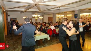 Eine Gruppe von Menschen tanzt in einem Bankettsaal. Es gibt Tische mit Tischdecken und Menschen, die um sie herum sitzen. Der Saal ist mit Kronleuchtern und einer Wandmalerei dekoriert.