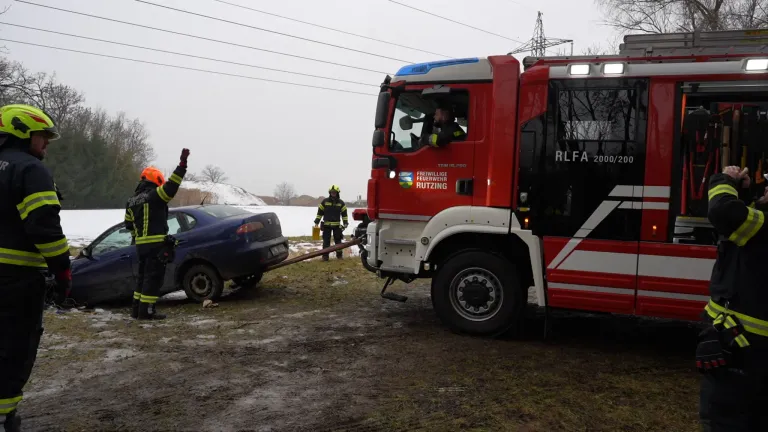 Ein blaues Auto wird von einem roten Feuerwehrauto gezogen. Ein Feuerwehrmann steht neben dem Auto.