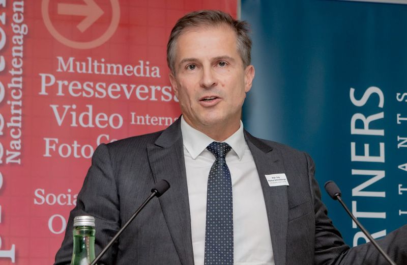 Ein Mann steht an einem Podium und hält eine Rede. Er trägt einen grauen Anzug und eine Krawatte. Hinter ihm befindet sich ein roter Banner mit dem Text Multimedia Presseverstoring Video International Foto und ein blaues Banner mit dem Wort Partners.