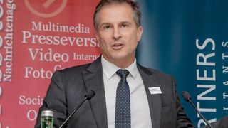 Ein Mann steht an einem Podium und hält eine Rede. Er trägt einen grauen Anzug und eine Krawatte. Hinter ihm befindet sich ein roter Banner mit dem Text Multimedia Presseverstoring Video International Foto und ein blaues Banner mit dem Wort Partners.