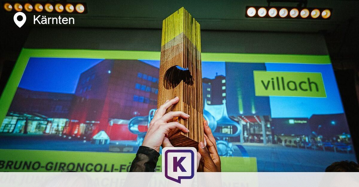 Zwei Hände halten einen Holzpreis vor einem Bildschirm, der ein Gebäude und das Wort 'Villo' zeigt. Ein K-Logo ist ebenfalls sichtbar.