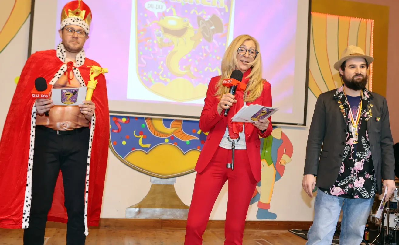 Eine Frau in einem roten Anzug hält ein Mikrofon und einen Schlüssel und steht vor einem farbenfrohen Hintergrund. Hinter ihr zeigt eine Leinwand einen animierten Charakter. In der Nähe stehen zwei Personen, eine in einem festlichen Kostüm und die andere in einem dunklen Anzug.