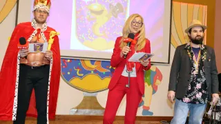 Eine Frau in einem roten Anzug hält ein Mikrofon und einen Schlüssel und steht vor einem farbenfrohen Hintergrund. Hinter ihr zeigt eine Leinwand einen animierten Charakter. In der Nähe stehen zwei Personen, eine in einem festlichen Kostüm und die andere in einem dunklen Anzug.