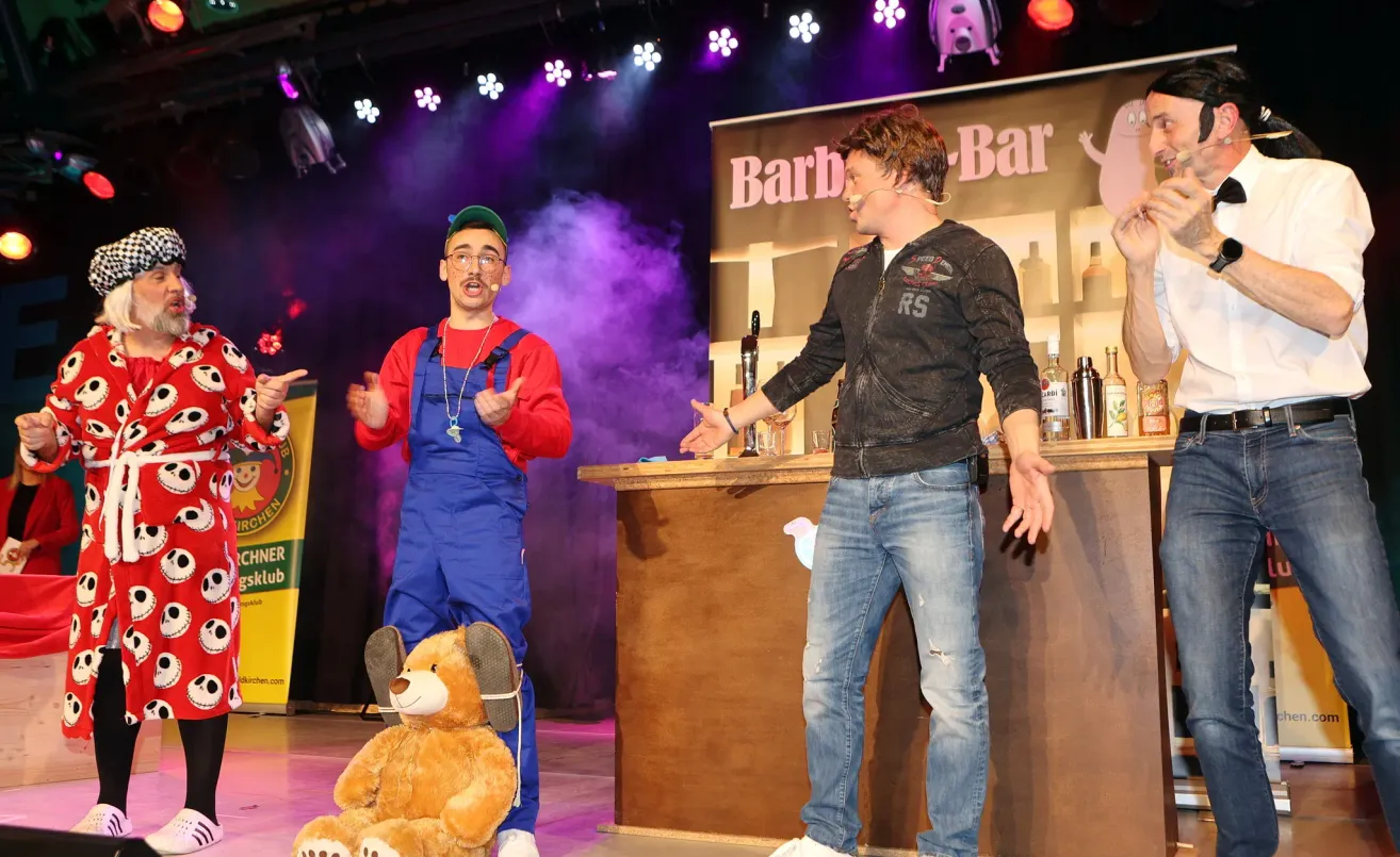 Zwei Männer auf der Bühne, einer als Mario gekleidet, sprechen zu einem Publikum. Dahinter steht eine hölzerne Bar mit Flaschen und einem Schild, das 'Barb-Bar' besagt. Ein Teddybär steht in der Nähe des Mario-Kostüms.