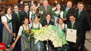 Eine Gruppe von Menschen in grüner und weißer traditioneller Kleidung posiert mit einer großen Karte einer Region, hält ein Zertifikat und Blumen. Ein Mann in einem grünen Anzug gibt einen Daumen hoch.