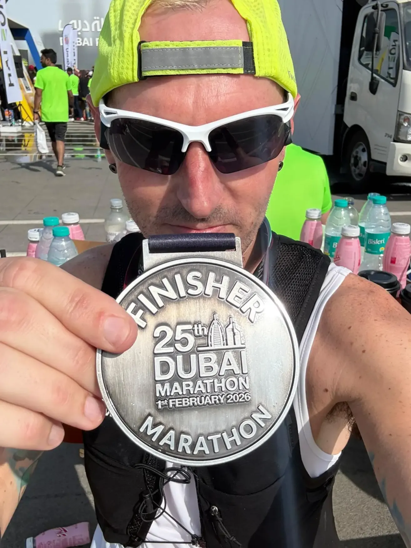 Ein Mann mit Sonnenbrille und gelber Mütze hält eine Finisher-Medaille für den 25. Dubai-Marathon, der am 1. Februar 2026 stattfand. Er steht vor einer Reihe von Flaschen und einem unscharfen Hintergrund.