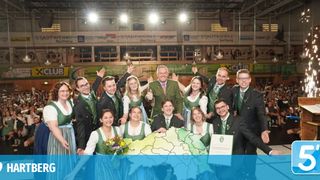 Eine Gruppe von Menschen in traditioneller deutscher Kleidung steht um eine Karte herum, einer hält ein Zertifikat. Sie posieren für ein Foto unter einem Scheinwerfer.