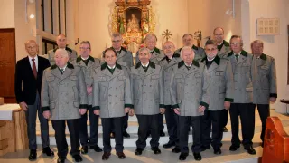 Ein Chor älterer Männer in grauen Mänteln mit grünem Besatz steht vor einem Kirchenaltar. Dahinter ist ein goldenes Altarbild mit einem religiösen Gemälde zu sehen.