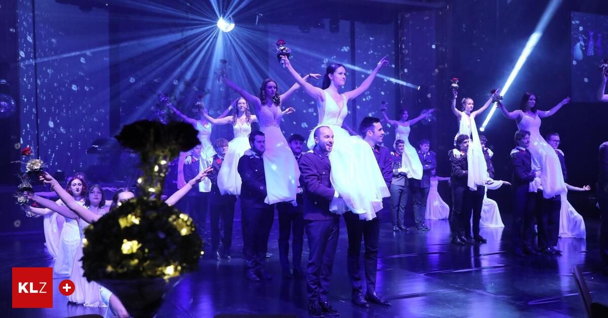 Eine Gruppe von Tänzern tritt auf einer Bühne unter violetten Lichtern auf. Sie halten rote Blumen und tragen weiße Kleider. Zwei Männer halten die Frauen in der Luft.