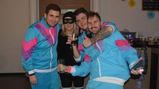 Vier Personen posieren für ein Foto, alle tragen passende blaue und rosa Trainingsanzüge. Einer hat ein Tattoo am Arm, und ein anderer hält ein Getränk. Eine Frau mit einer Maske steht hinter ihnen.