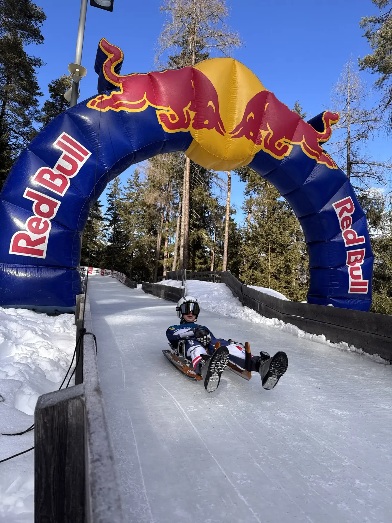 Eine Person mit Helm fährt auf einem Schlitten die Eisbahn hinunter, mit einem Red-Bull-Bogen im Hintergrund.