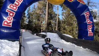Eine Person mit Helm fährt auf einem Schlitten die Eisbahn hinunter, mit einem Red-Bull-Bogen im Hintergrund.