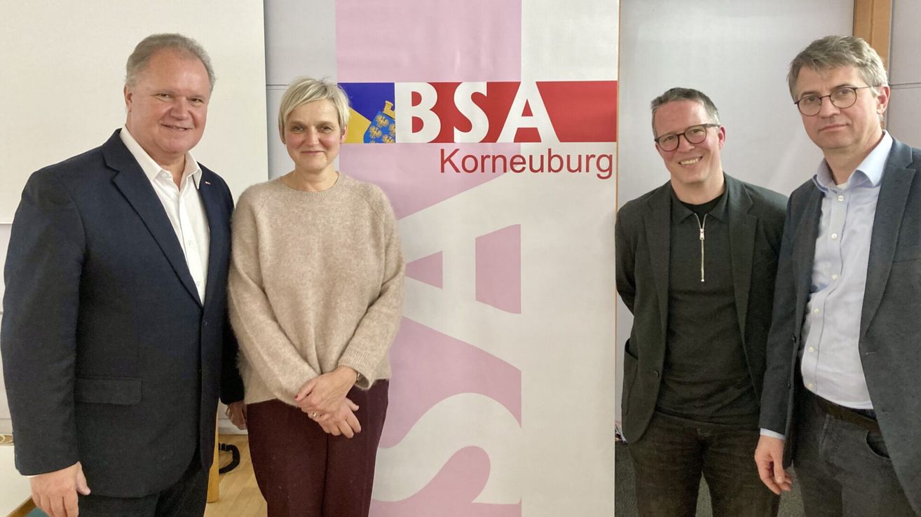 Drei Personen stehen vor einem Banner mit dem Text BSA Korneuburg. Eine Frau in einem beigen Pullover und einer braunen Hose steht zwischen zwei Männern, einem in einem Anzug und der andere in einem schwarzen Hemd und einer Brille.