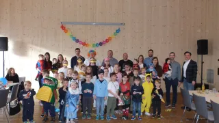 Eine Gruppenaufnahme von Kindern und Erwachsenen in Kostümen, die in einem Raum mit Konfetti, Holzwänden und einem Banner aus bunten Blumen stehen.