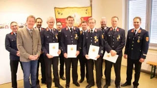 Eine Gruppe von Männern in Uniform, möglicherweise Feuerwehrleute, stehen in einer Reihe, halten Urkunden und lächeln für ein Foto in einem Raum mit einem Banner.