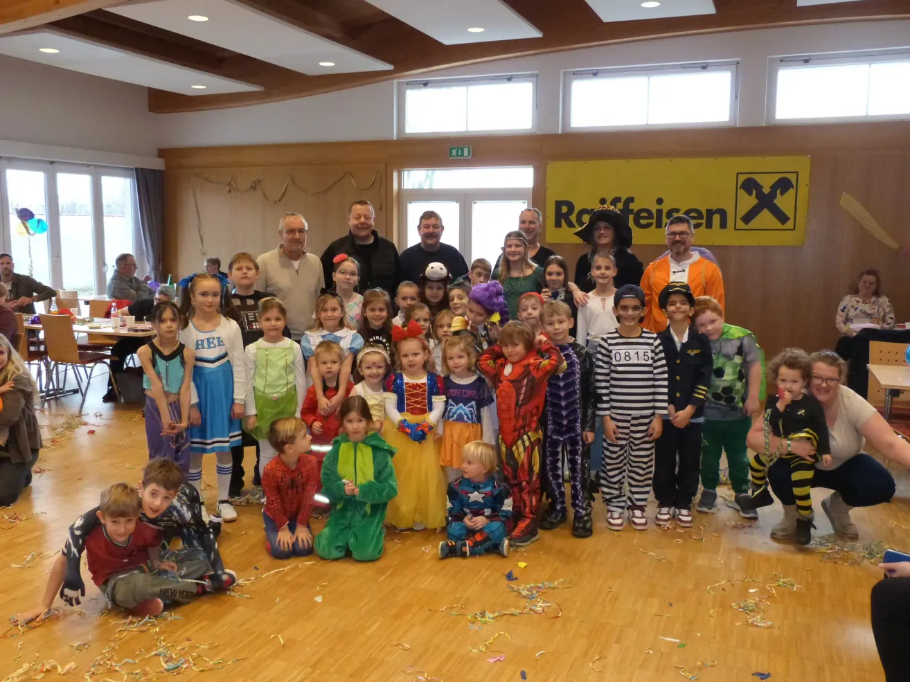 Eine Gruppe von Kindern und Erwachsenen, die verschiedene Kostüme tragen, posiert für ein Foto in einem Raum. Hinter ihnen ist ein gelbes Banner mit dem Wort 'Reifen' zu sehen.