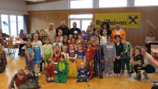 Eine Gruppe von Kindern und Erwachsenen, die verschiedene Kostüme tragen, posiert für ein Foto in einem Raum. Hinter ihnen ist ein gelbes Banner mit dem Wort 'Reifen' zu sehen.