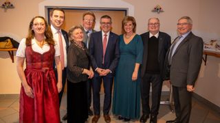 Eine Gruppe formell gekleideter Personen posiert für ein Foto, steht eng beieinander und lächelt, mit einer Holztür und Kunstwerk im Hintergrund.