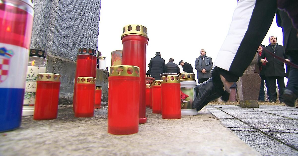Eine Gruppe von Menschen versammelt sich um ein Denkmal mit roten Kerzen und einer weißen Urne, möglicherweise bei einer Beerdigung.