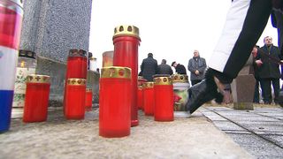 Eine Gruppe von Menschen versammelt sich um ein Denkmal mit roten Kerzen und einer weißen Urne, möglicherweise bei einer Beerdigung.