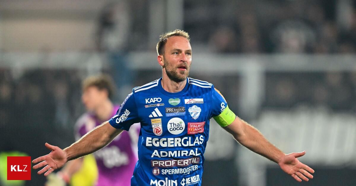 Ein Fußballspieler in einem blauen Trikot feiert. Seine Arme sind weit ausgebreitet. Dahinter beobachtet ein anderer Spieler in einem lila Trikot.